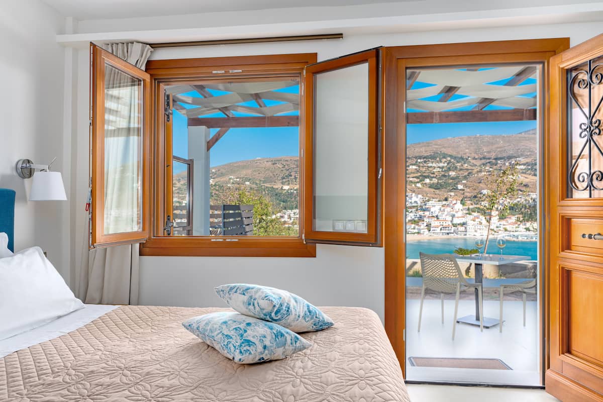 A bedroom with a bed, two pillows, a nightstand, a lamp, and a window with a view of a coastal town.