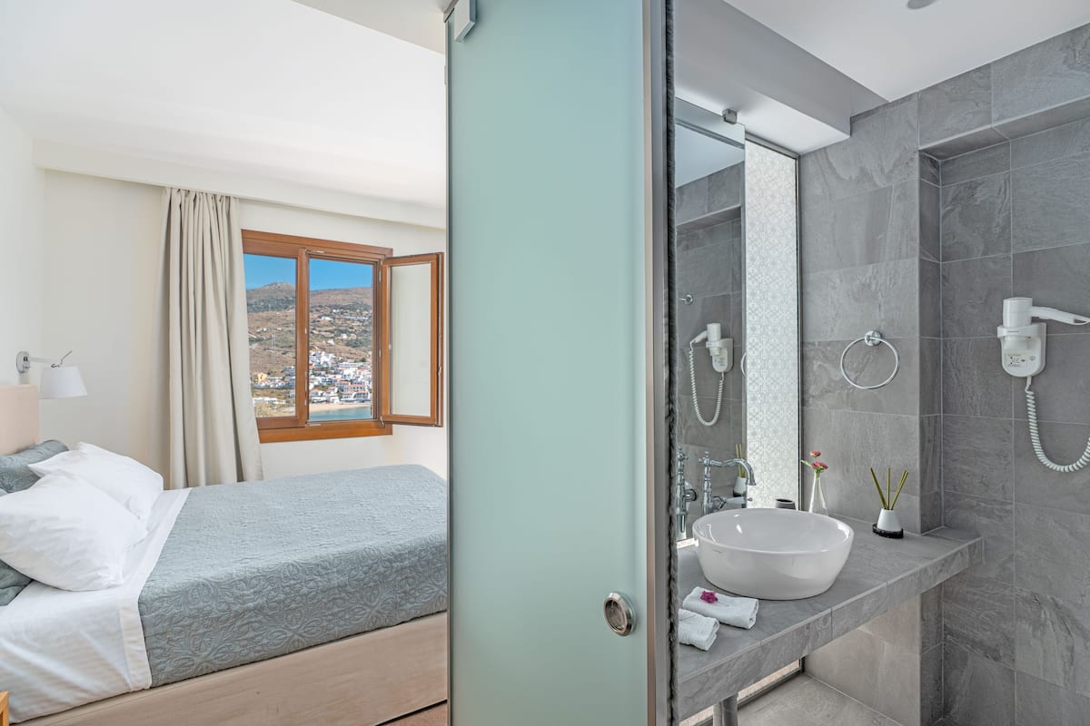 A modern hotel room with a bed, a bathroom with a large mirror, a sink, and a view of a coastal town.