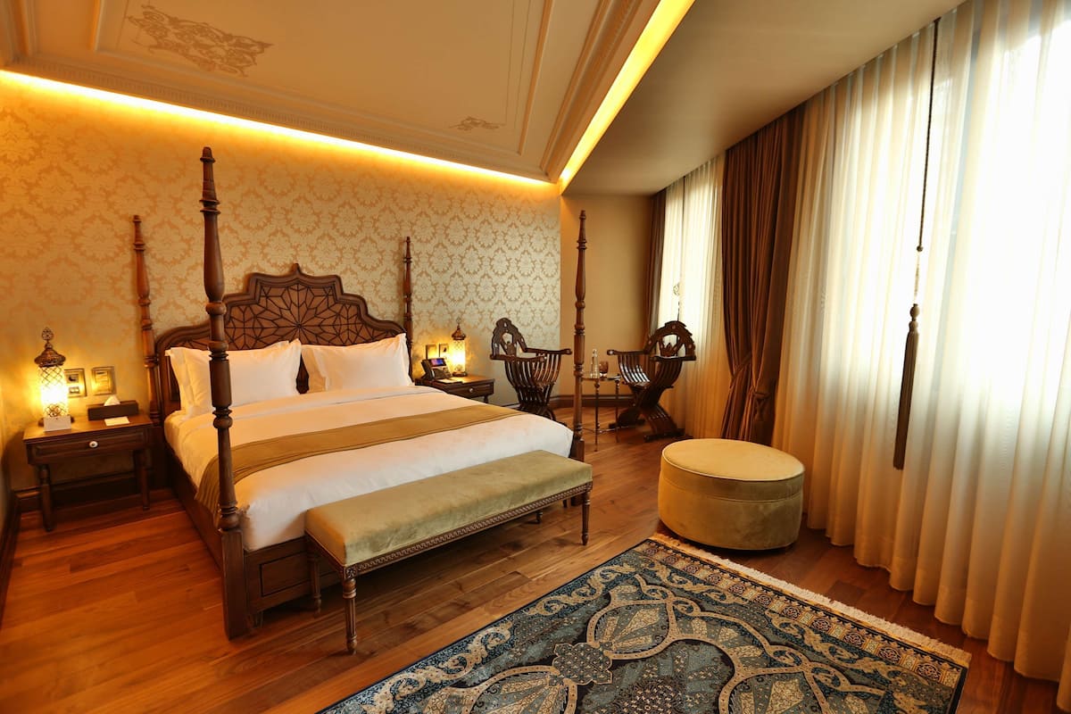 A four-poster bed with a bench, a wooden floor, a patterned rug, and a large window with curtains.