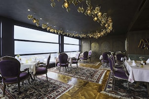 A dining area with tables set for six, purple upholstered chairs, and a large view of the city through large windows.