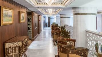 A grand hallway with ornate chandeliers, wooden paneling, and a series of chairs with intricate designs.