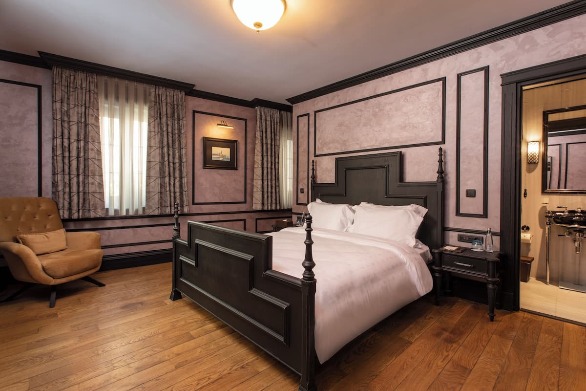 A bedroom with a four-poster bed, a nightstand, a chair, and a bathroom with a sink and mirror.