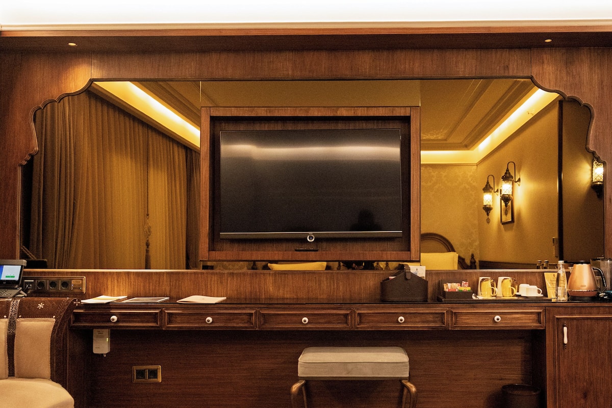 A hotel room with a large flat-screen TV, wooden furniture, a bench, a desk, and a patterned rug.