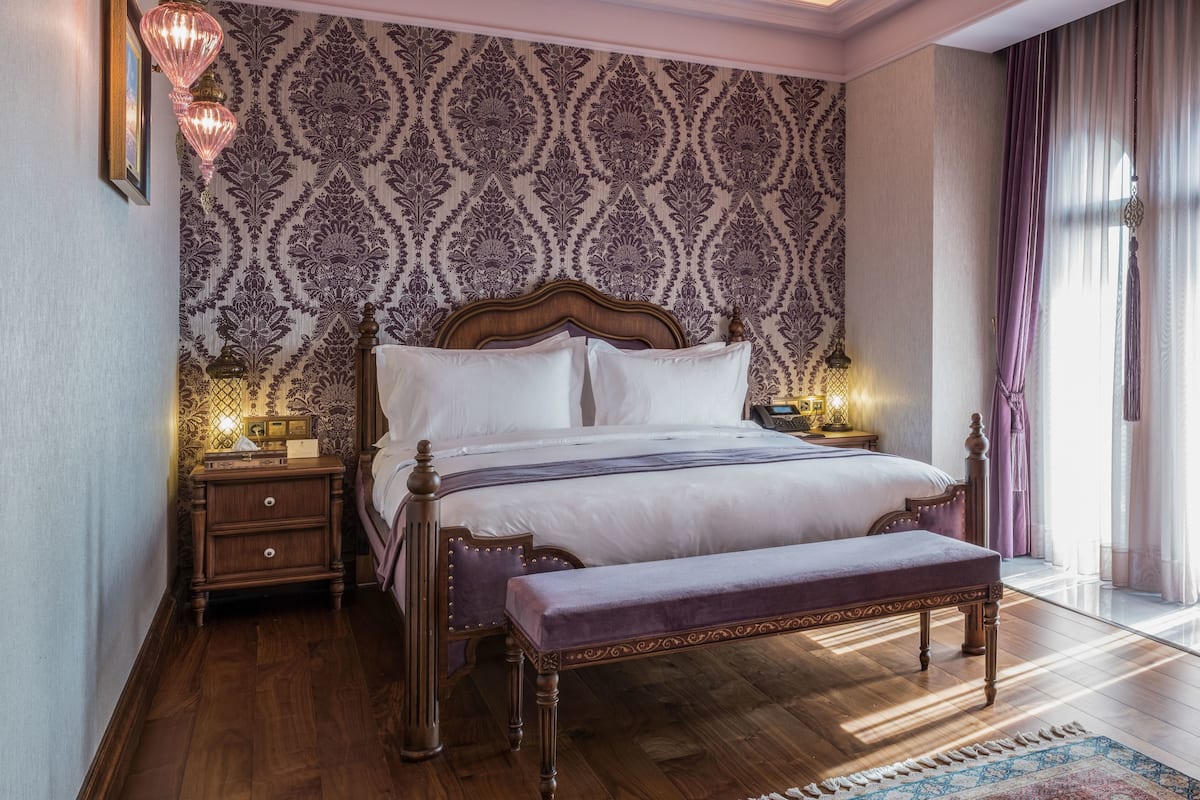 A bedroom with a large bed, wooden headboard, and a bench. There is a nightstand with a lamp and a small table with a lamp. The room has patterned wallpaper and a wooden floor.