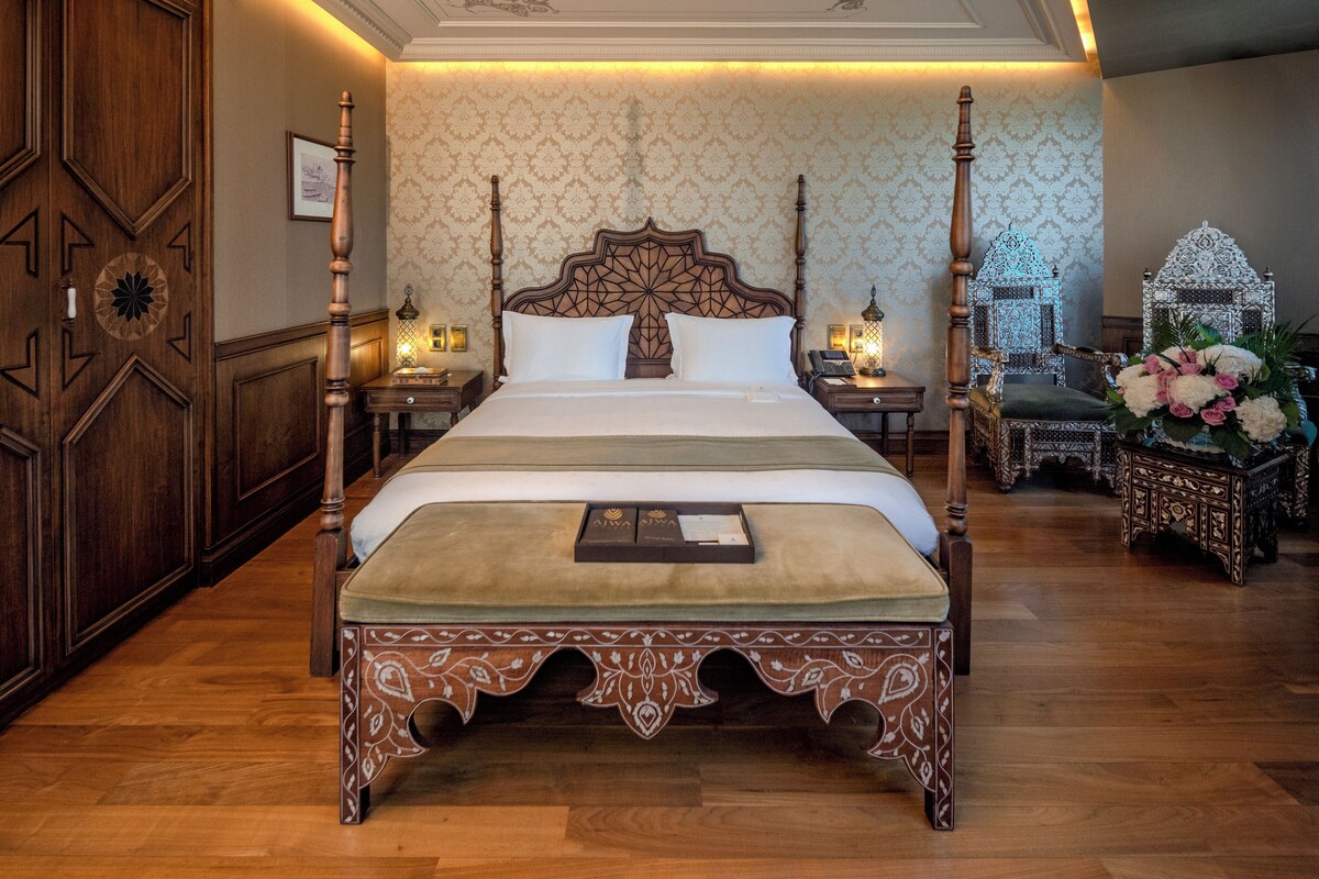 A four-poster bed with a decorative headboard, a nightstand with a lamp, and a floral arrangement on the right.