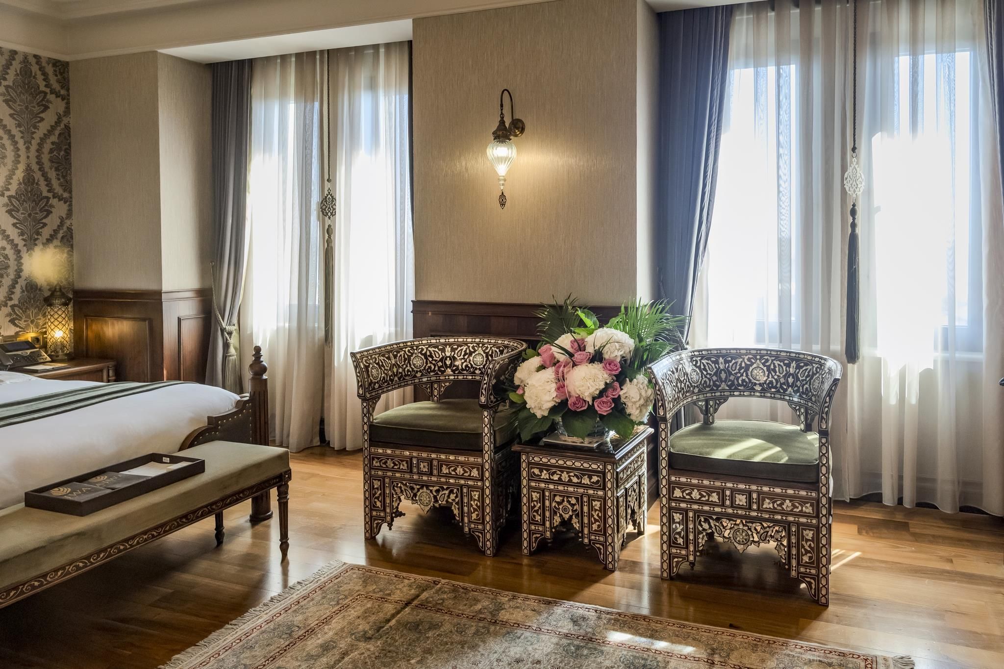 A hotel room with a bed, two ornate chairs, a small table with a floral arrangement, and a wall-mounted lamp.