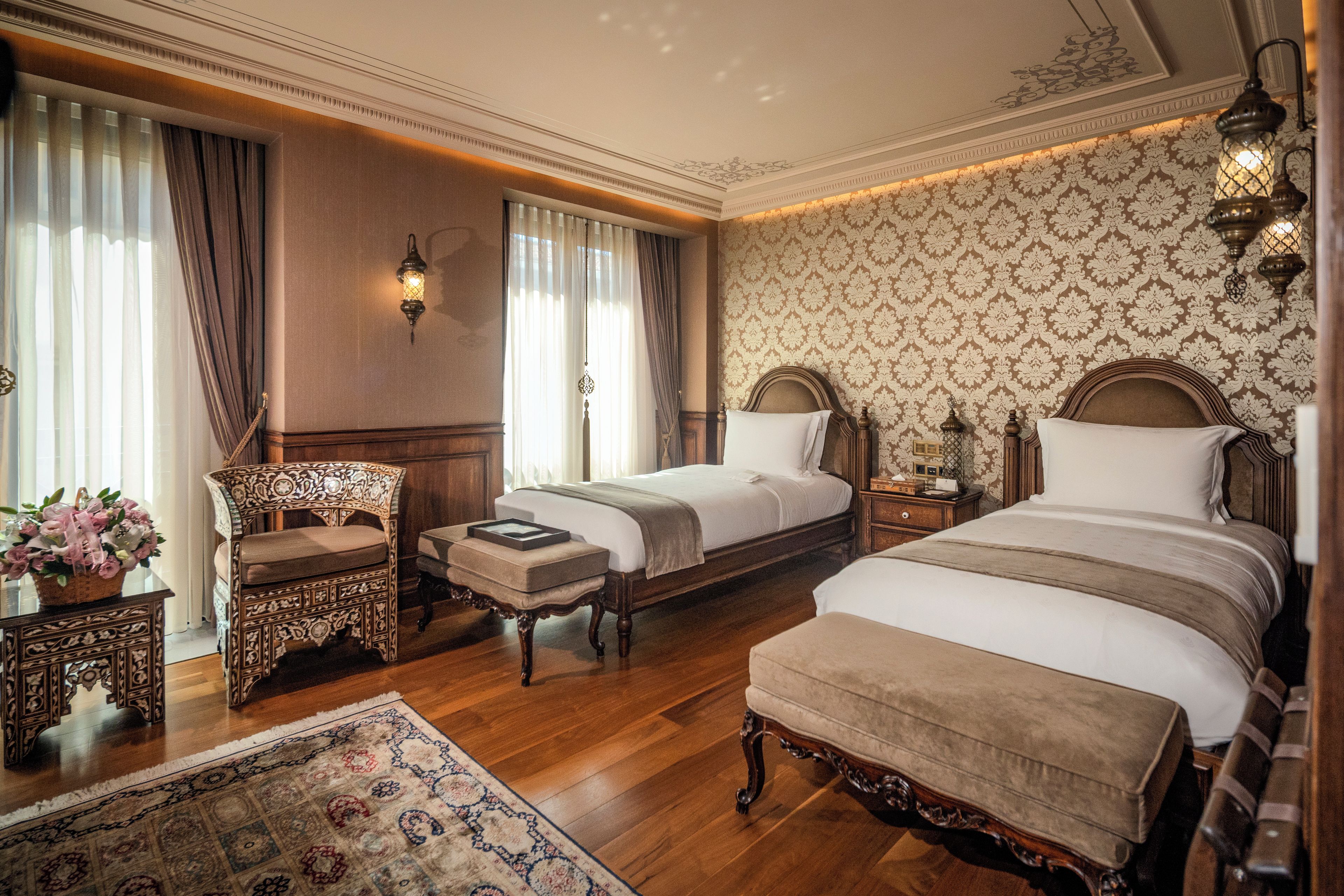 A hotel room with two beds, a sitting area with a chair and table, a decorative rug, and a patterned wallpaper.