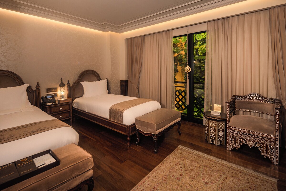 A hotel room with two beds, a sitting area with a chair, a small table, and a view of greenery through the window.