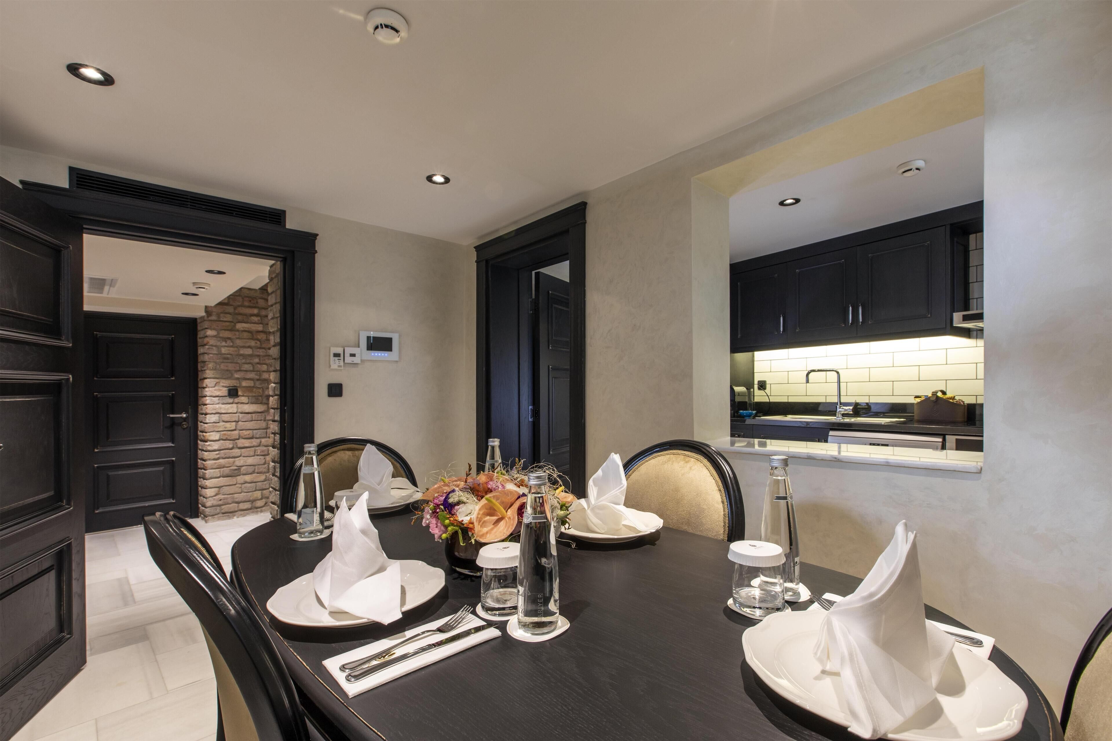 A dining area with a dark wooden table set for four, surrounded by dark wooden chairs. There is a decorative centerpiece on the table, and the room has a modern kitchen visible through an open doorway.