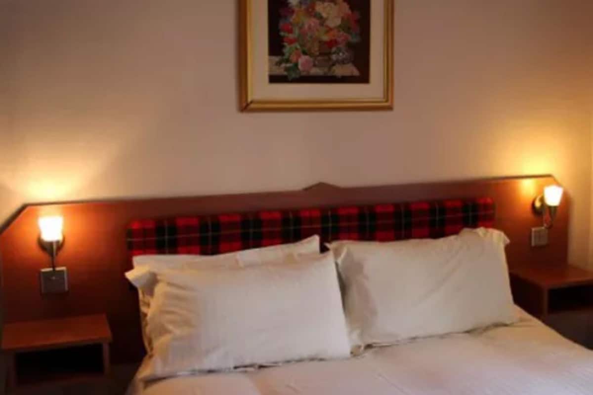 A bed with white linens, flanked by two bedside tables with lamps, a red plaid headboard, and a framed floral artwork on the wall.