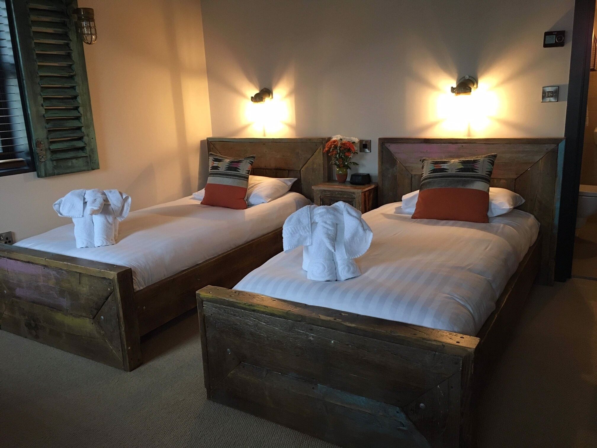 Dos camas de madera con sábanas blancas, cada una con una escultura de toalla, una mesita de noche, un jarrón con flores e iluminación montada en la pared.