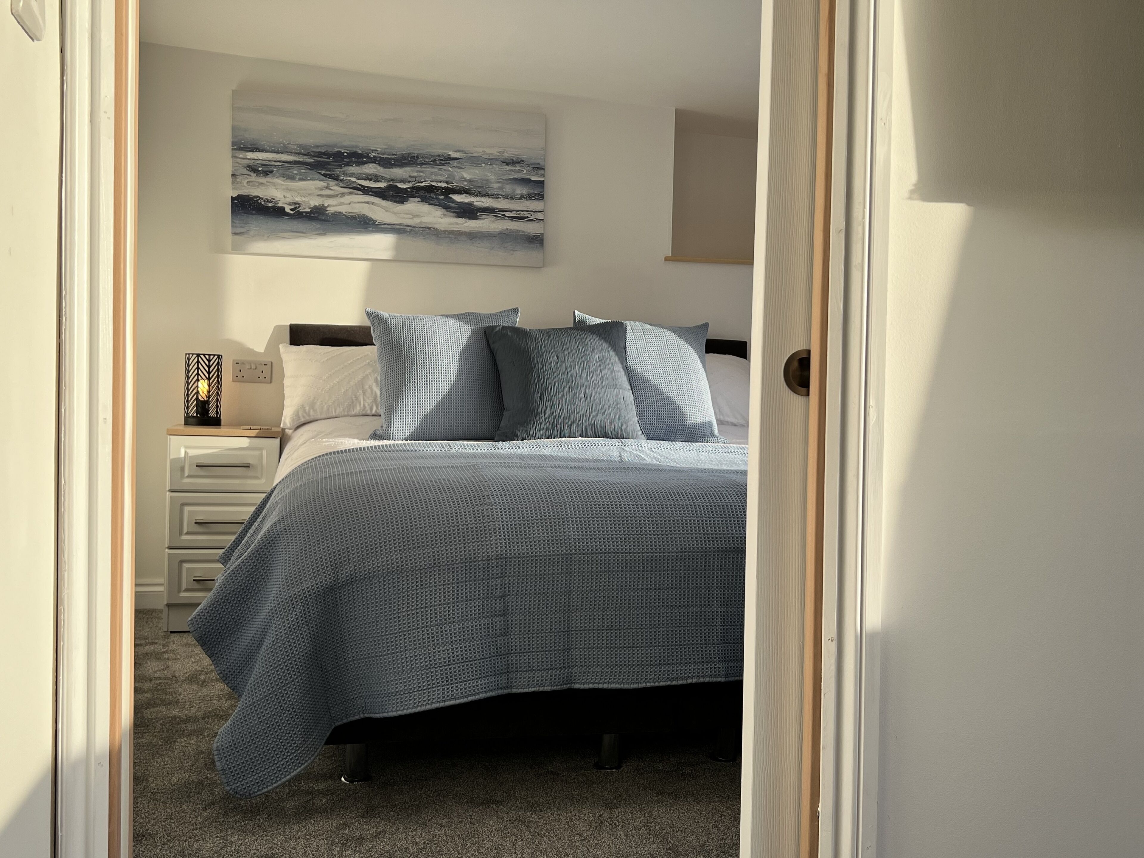 Un dormitorio con cama, almohadas, una mesita de noche, lámpara y un cuadro en la pared.