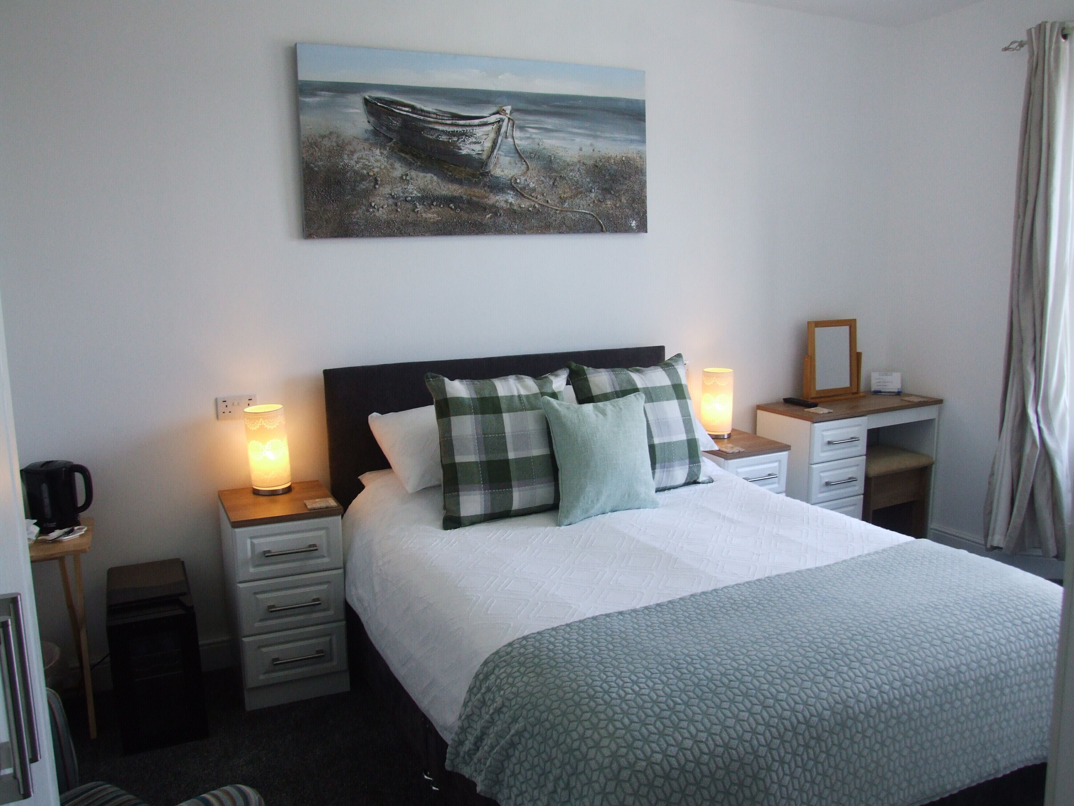 A neatly made bed with a checkered pillow, a bedside lamp, and a painting of a boat on the wall.