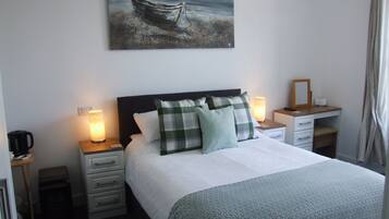A neatly made bed with a checkered pillow, a bedside lamp, and a painting of a boat on the wall.