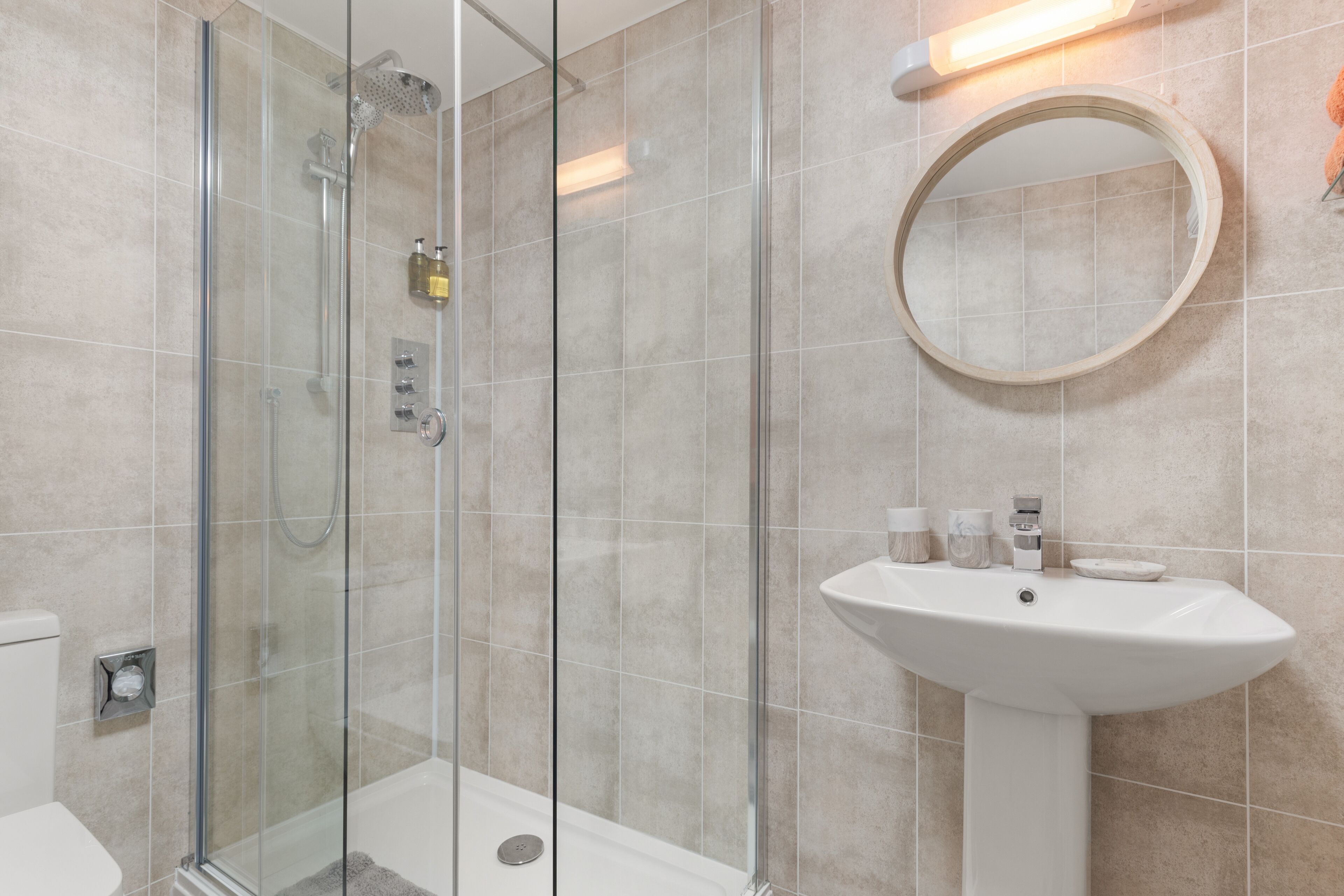 Un baño moderno con ducha envuelta en vidrio, lavamanos sobre pedestal y espejo redondo.