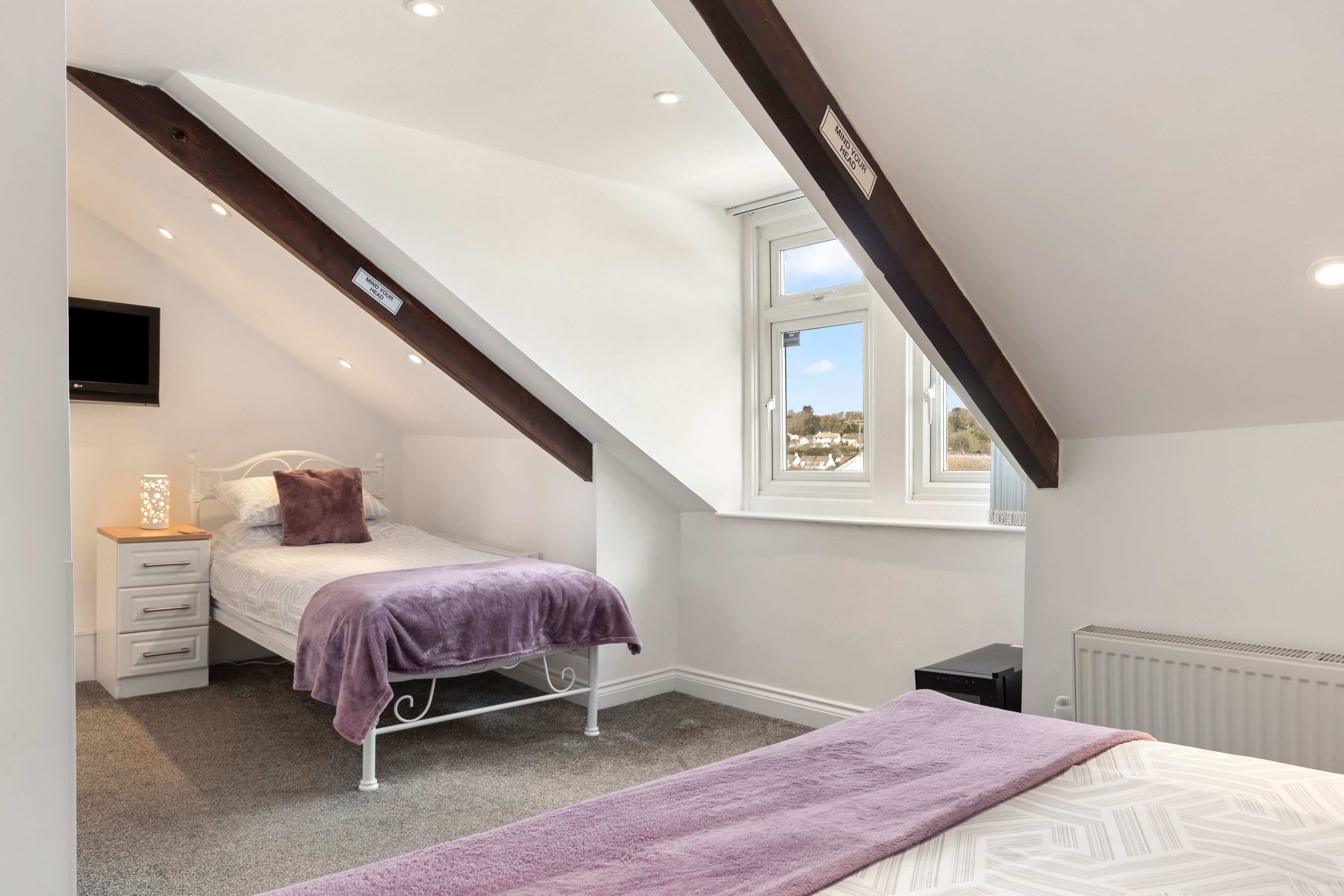 A bedroom with a sloped ceiling, a bed with a purple blanket, a bedside table with a lamp, and a window with a view.