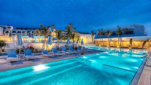 Une piscine entourée de palmiers et d’immeubles, avec des chaises longues et des parasols.