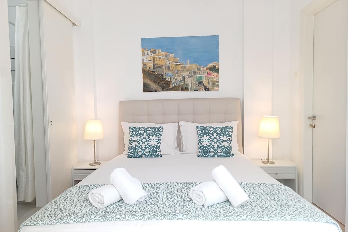 Una cama bien hecha con ropa de cama estampada, dos lámparas de noche y un cuadro de un pueblo costero en la pared.