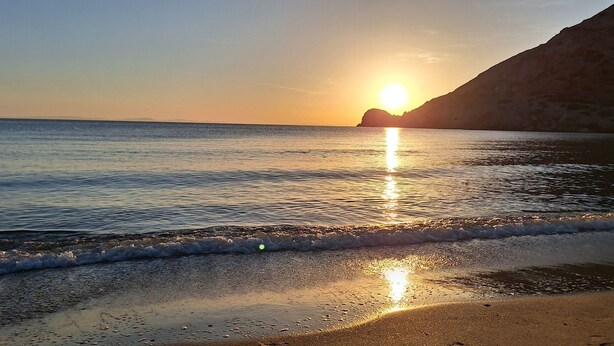 Une scène de plage avec un coucher de soleil sur l’océan, des vagues qui viennent doucement lécher le rivage et une falaise en arrière-plan.