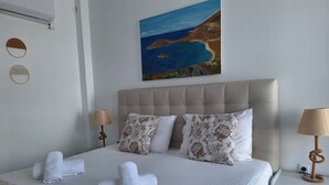 Un lit bien fait, avec du linge de lit blanc et des coussins décoratifs, un tableau représentant un paysage côtier au-dessus du lit, et une table de chevet avec une lampe.