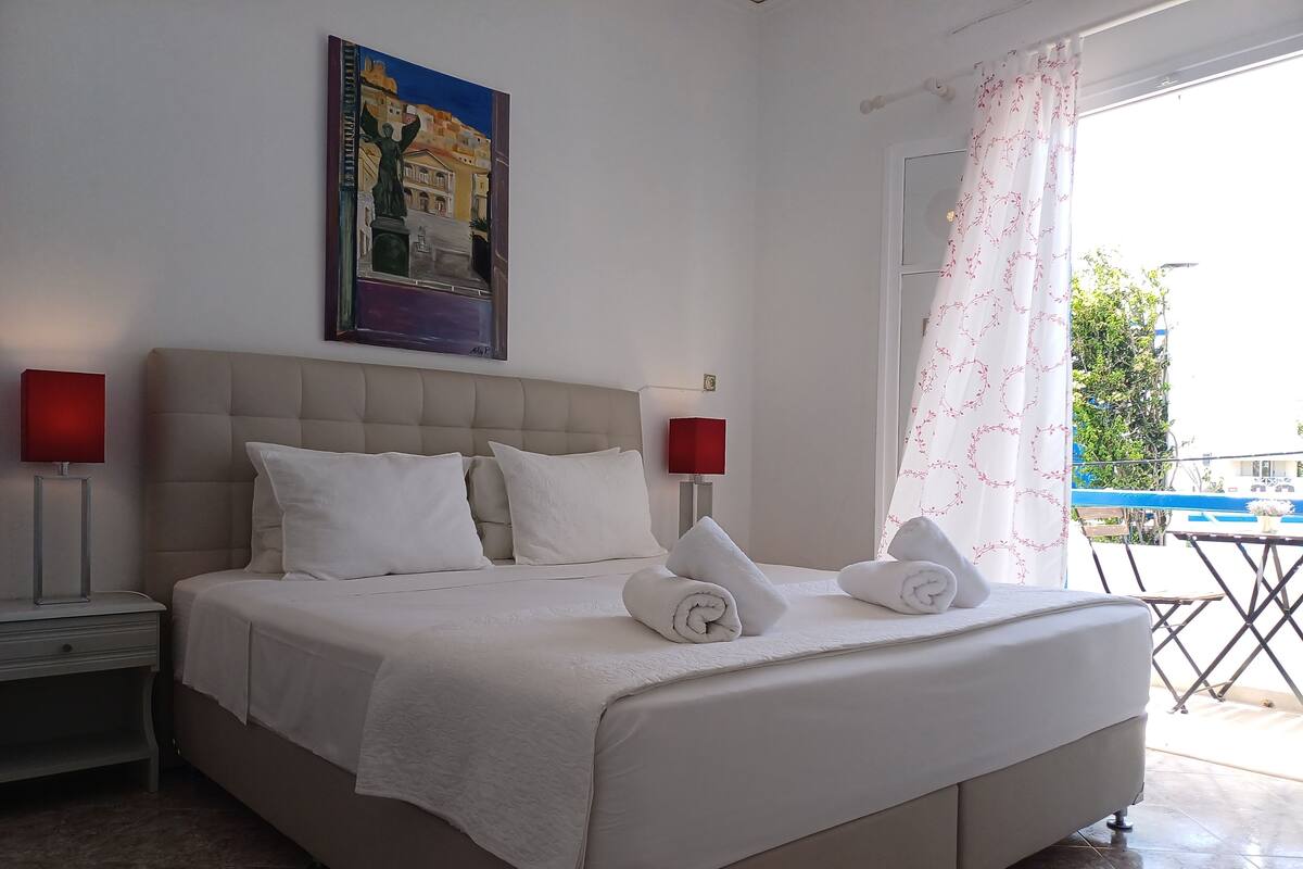 Una cama bien hecha con sábanas blancas y dos toallas enrolladas, un cabecero, dos lámparas de noche, un cuadro sobre la cama y vistas del exterior a través de una ventana con cortina blanca.