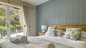 A bedroom with a large bed, a nightstand, a lamp, and a view of greenery through a window. - The Manor at Abberley (Worcester)