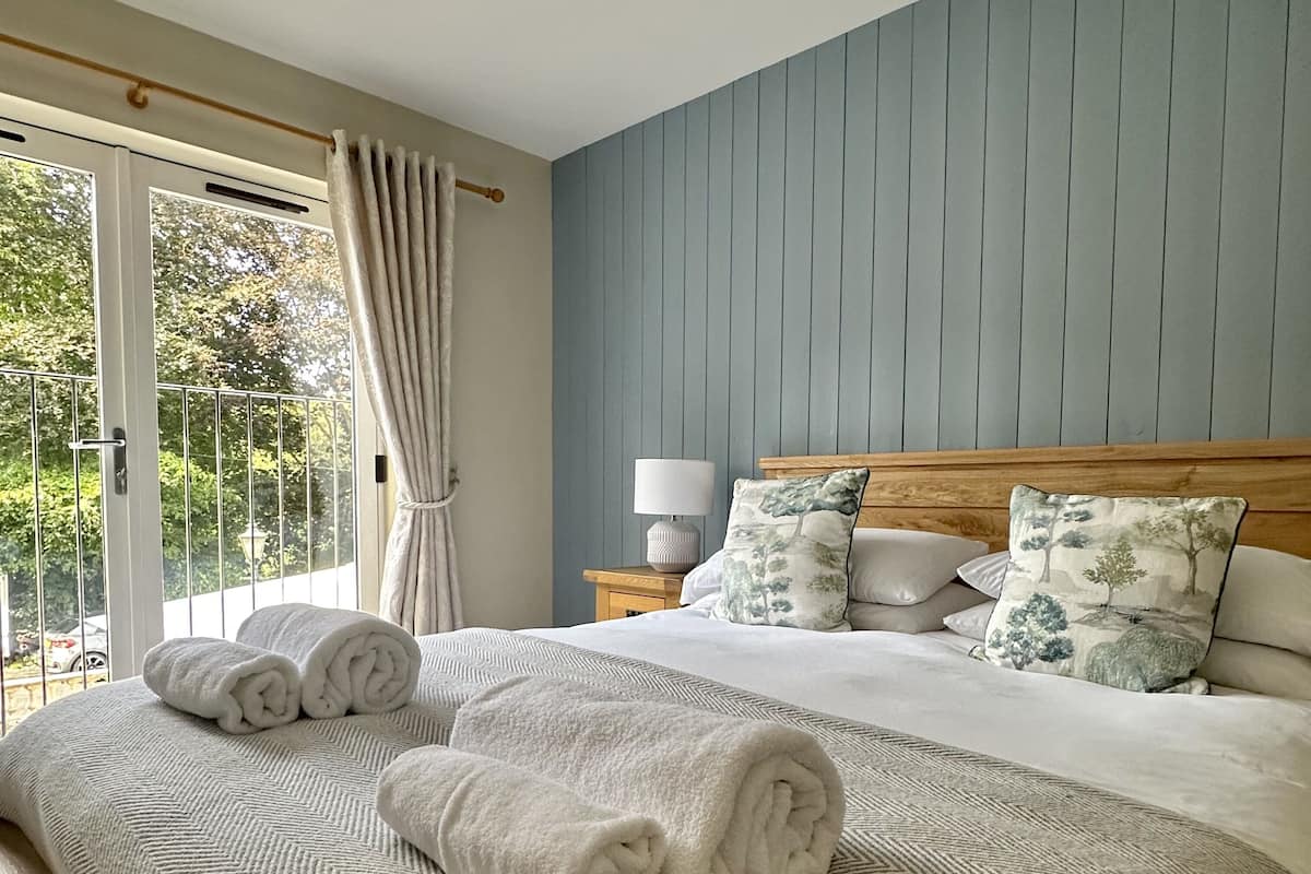 A bedroom with a large bed, a nightstand, a lamp, and a view of greenery through a window.