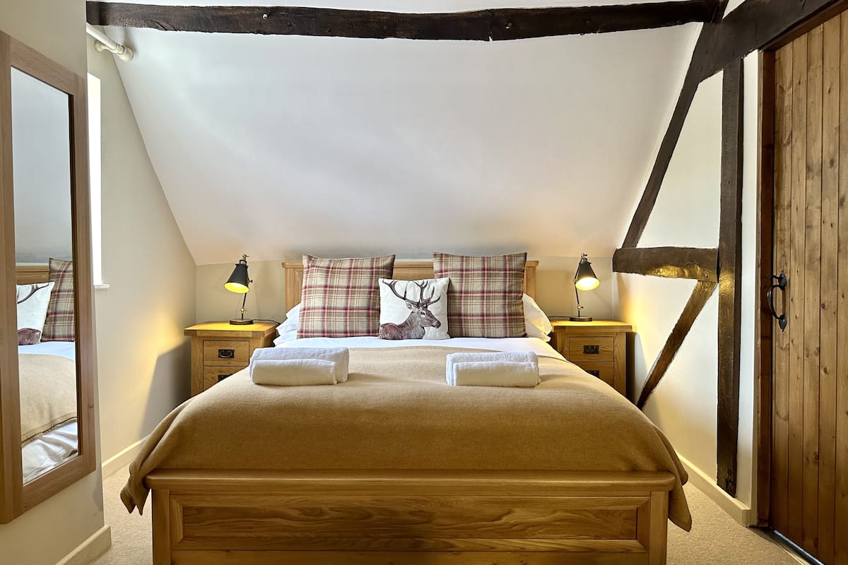 A cozy bedroom with a wooden bed, plaid pillows, and a deer-themed pillow. There are two bedside tables with lamps, and a wooden door with a metal handle.