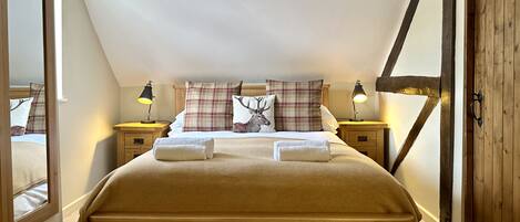 A cozy bedroom with a wooden bed, plaid pillows, and a deer-themed pillow. There are two bedside tables with lamps, and a wooden door with a metal handle.