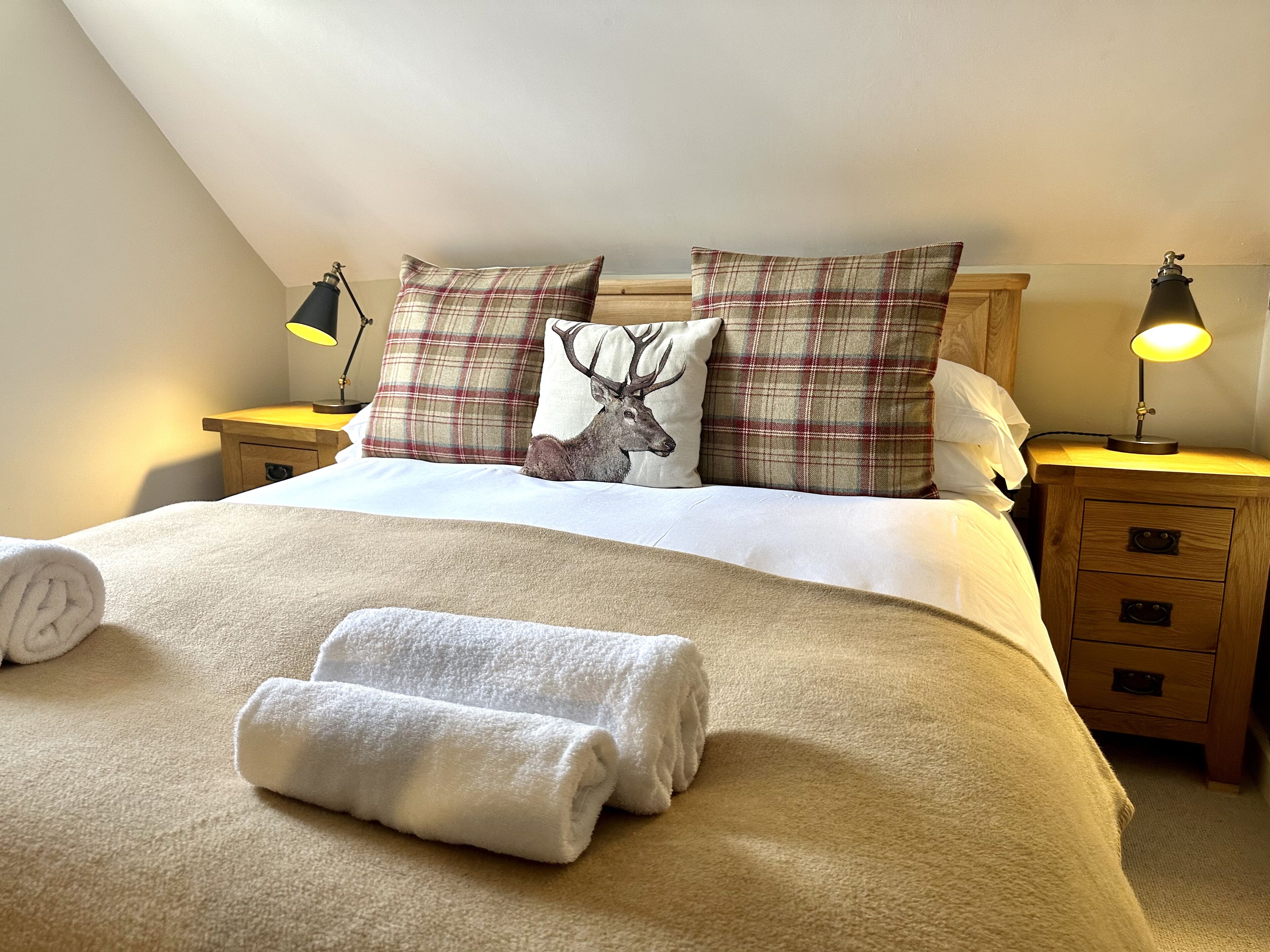 A neatly made bed with plaid pillows, a rolled white towel, and a nightstand with a lamp.