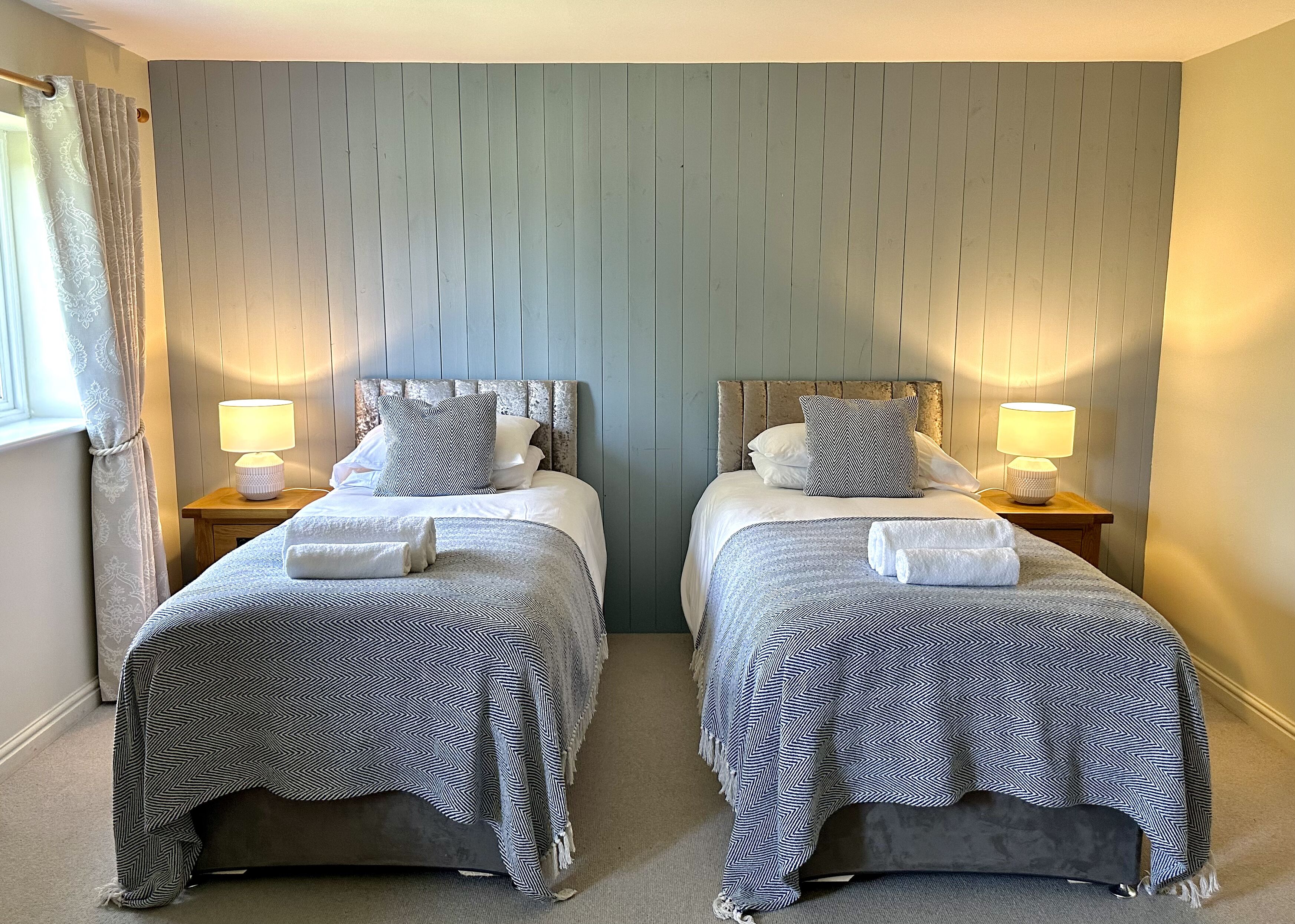 A room with two beds, each with a headboard, bedside lamps, and towels.