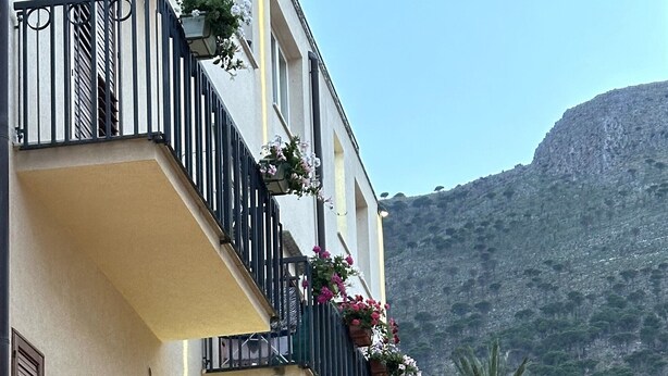 A multi-story building with balconies adorned with flowers, a stone wall, and a mountain in the background. - Badia Rooms & Breakfast (Castellammare del Golfo)