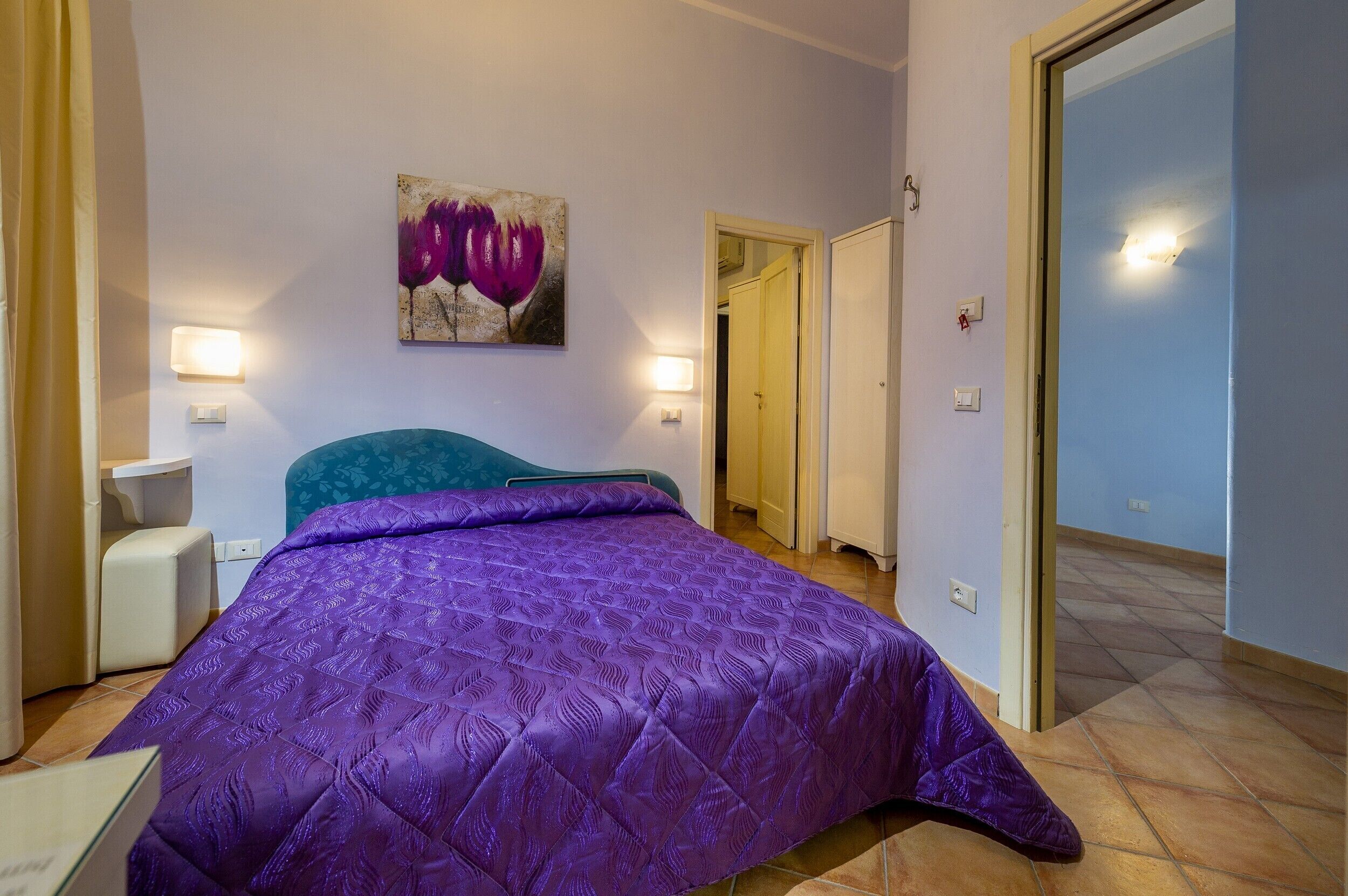 Un dormitorio con una colcha morada, un cuadro colgado en la pared, lámparas de noche y una puerta que da a otra habitación.