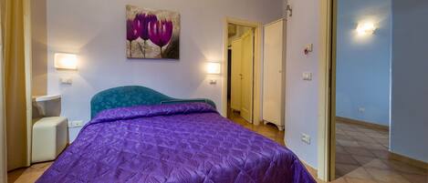 Un dormitorio con una colcha morada, un cuadro colgado en la pared, lámparas de noche y una puerta que da a otra habitación.