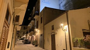 A narrow street with buildings on either side, a sign for "Bastia Rooms" on the right, and parked cars in the distance. - Badia Rooms & Breakfast (Castellammare del Golfo)