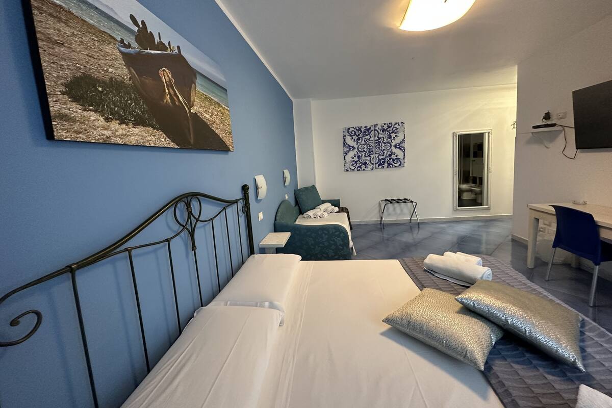 Une chambre d’hôtel comprenant un lit, un bureau, une chaise, une petite table, un téléviseur fixé au mur et un tableau représentant un bateau accroché au mur.