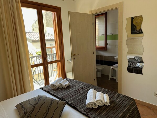 A hotel room with a bed, two towels, a bathroom with a toilet, and a view of the outside balcony.