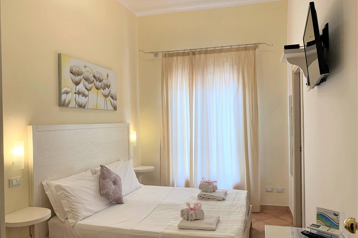 Une chambre d’hôtel avec un lit, une table de chevet, un téléviseur fixé au mur et une fenêtre avec des rideaux.