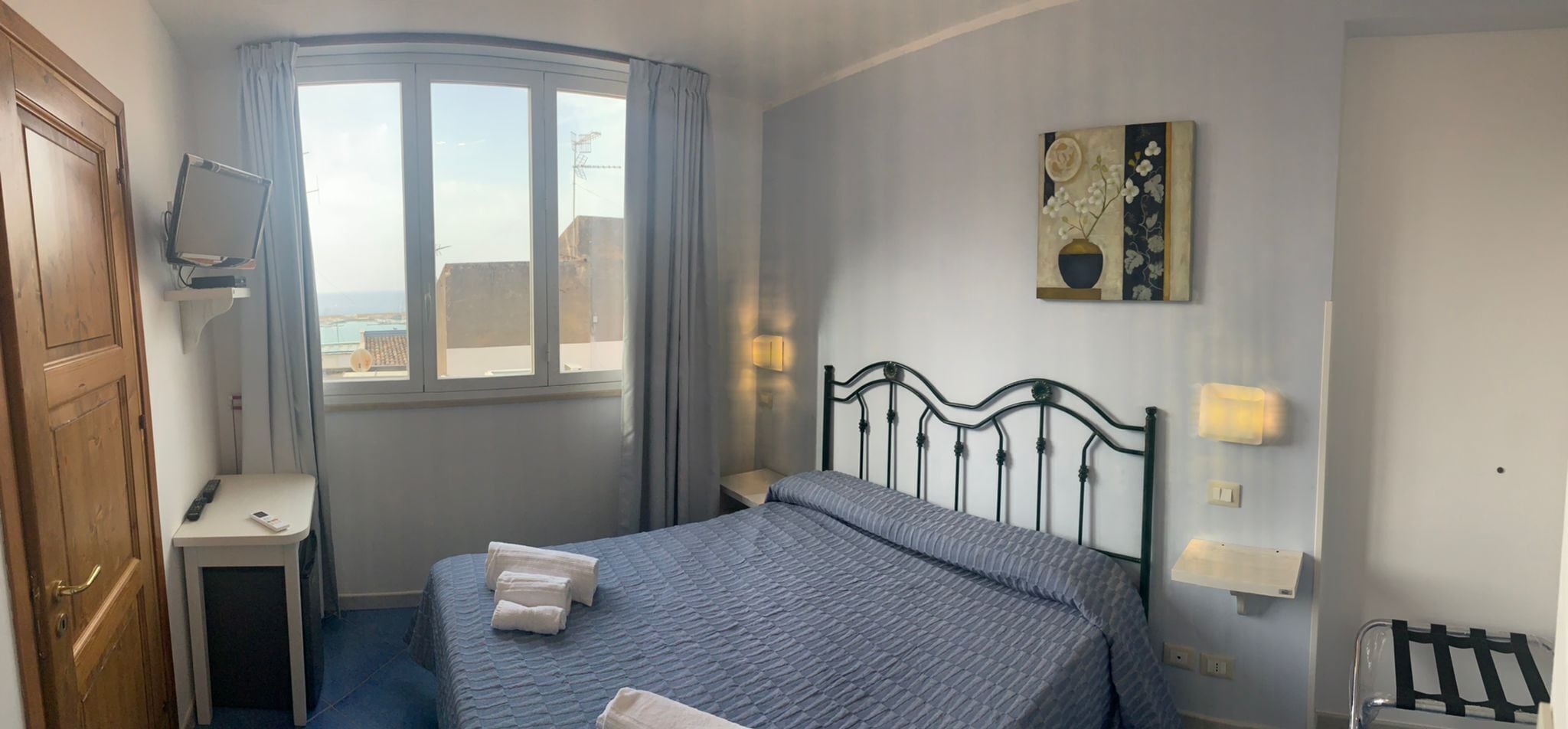 Ein Schlafzimmer mit Bett, Nachttisch, Fernseher, einem Fenster mit Vorhängen und einem Bild an der Wand.
