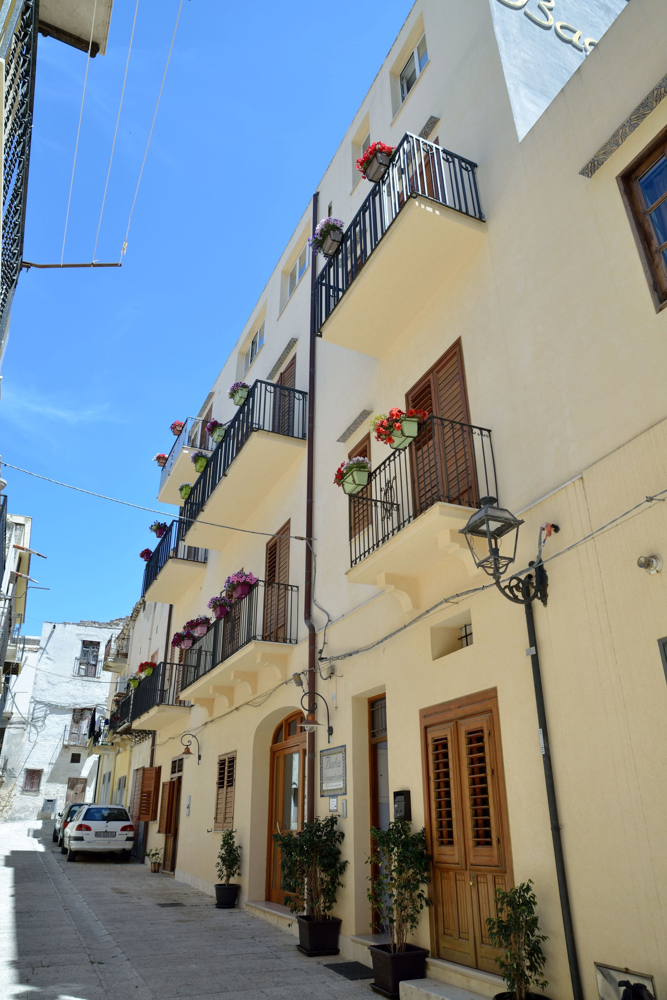 A narrow street with buildings featuring balconies with flower boxes, wooden doors, and a street lamp.