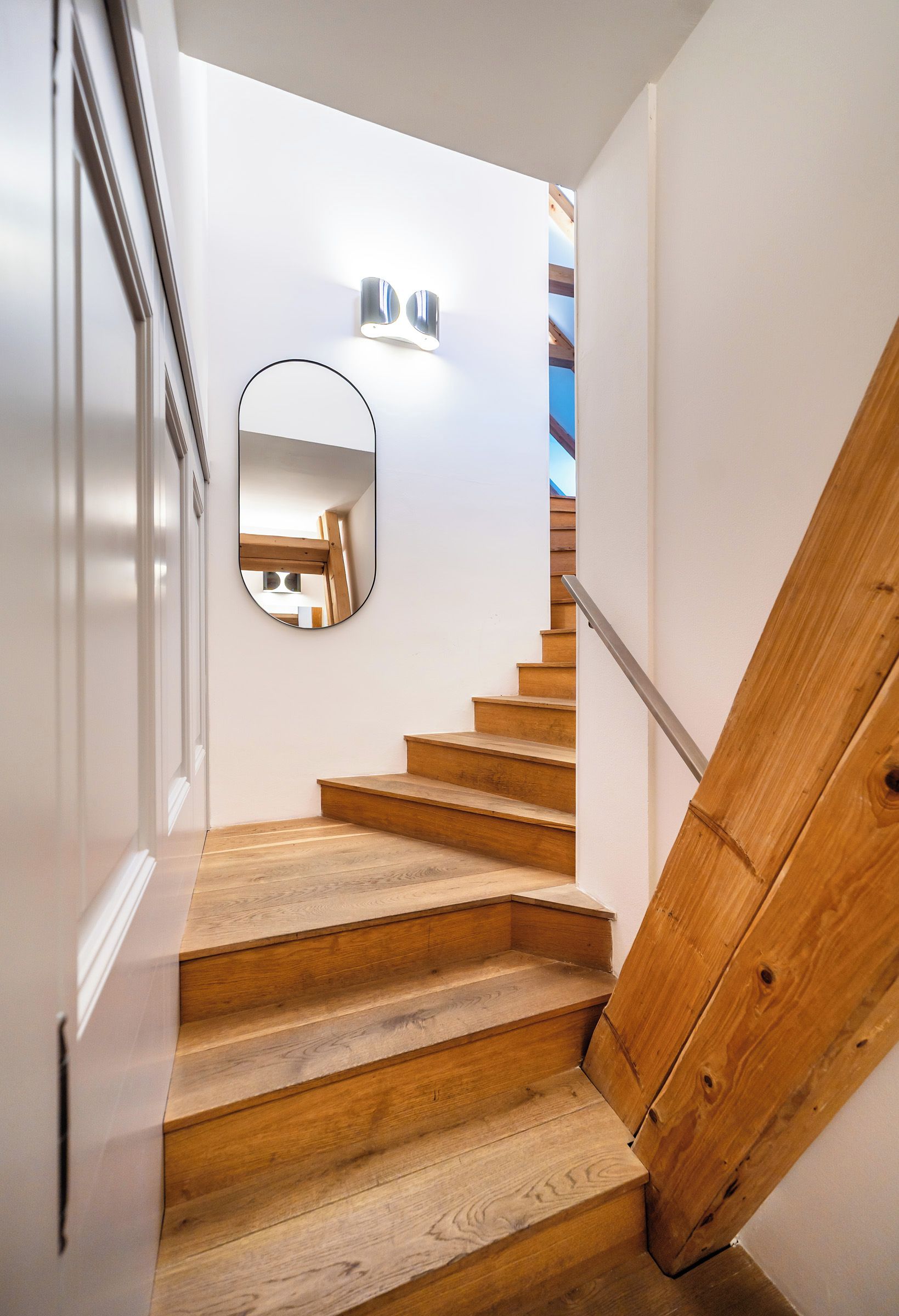 Una escalera de madera con barandilla, un espejo en la pared y una lámpara arriba.