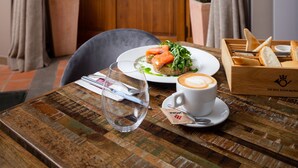 A dining area with a wooden table, a plate of food, a cup of coffee, and a window with curtains. - Old Royal Post Boutique Hotel & Premium Suites (Prague)