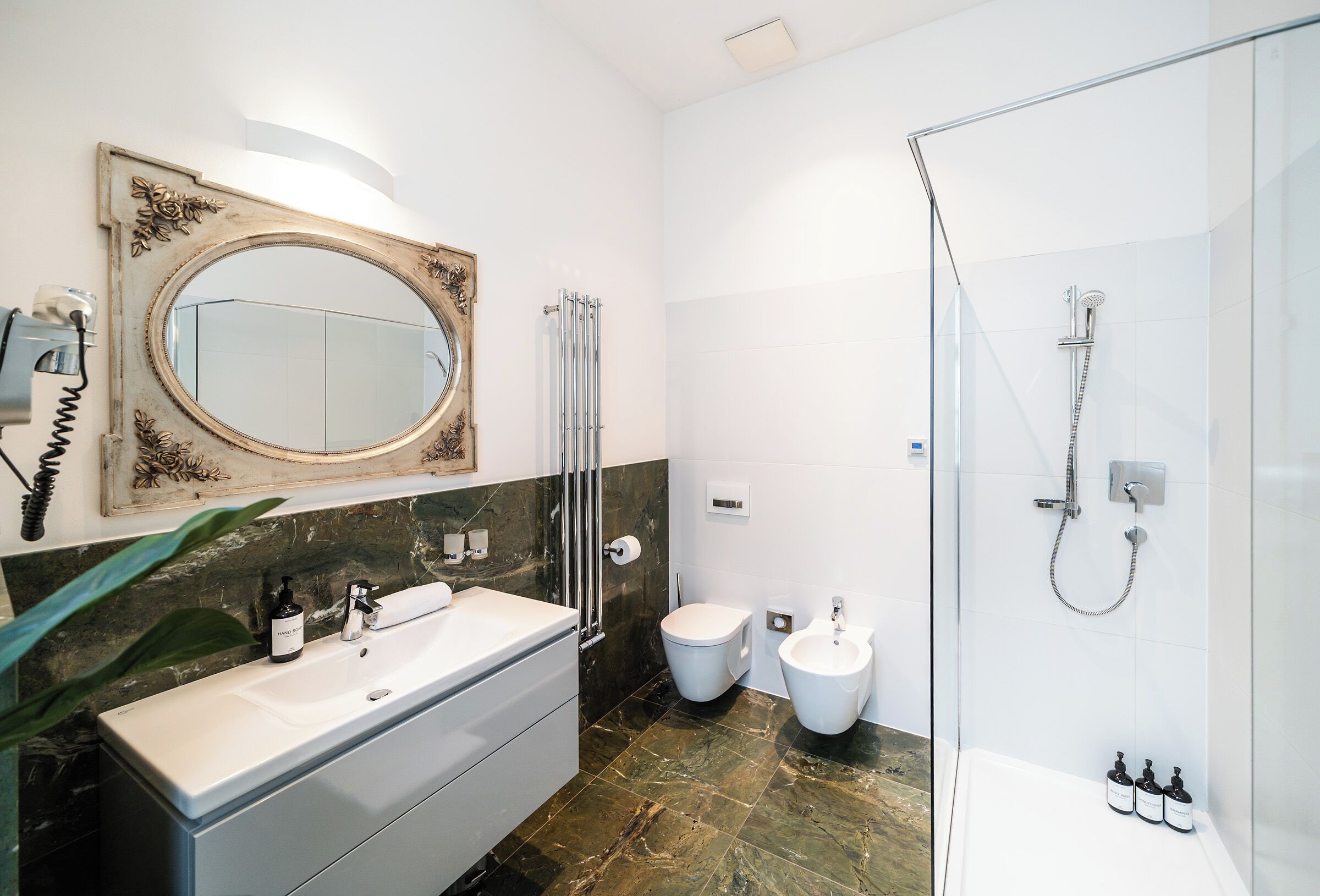 A modern bathroom with a large oval mirror, a white sink, a bidet, and a glass-enclosed shower area.