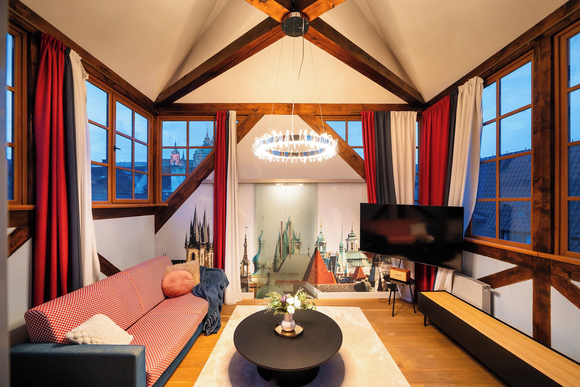 A cozy living room with a high ceiling, large windows, a flat-screen TV, and a chandelier.