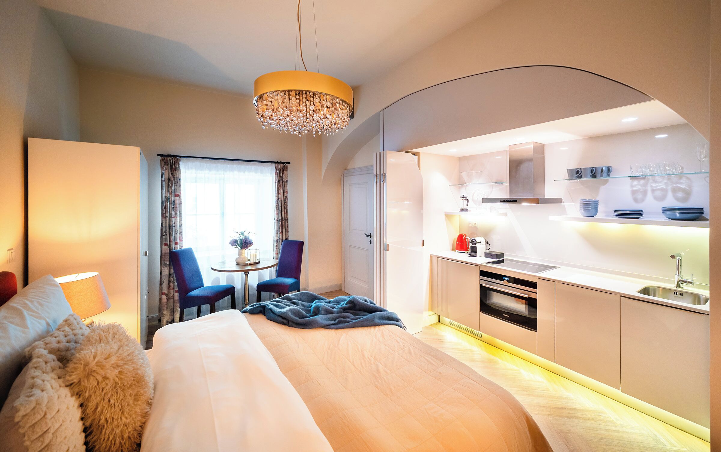 Ein modernes Hotelzimmer mit einem großen Bett, einer Sitzecke mit Sesseln, einem kleinen Tisch mit einer Vase, einer Küchenzeile mit Spüle und Geräten sowie einem Kronleuchter.