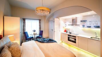 Ein modernes Hotelzimmer mit einem großen Bett, einer Sitzecke mit Sesseln, einem kleinen Tisch mit einer Vase, einer Küchenzeile mit Spüle und Geräten sowie einem Kronleuchter.