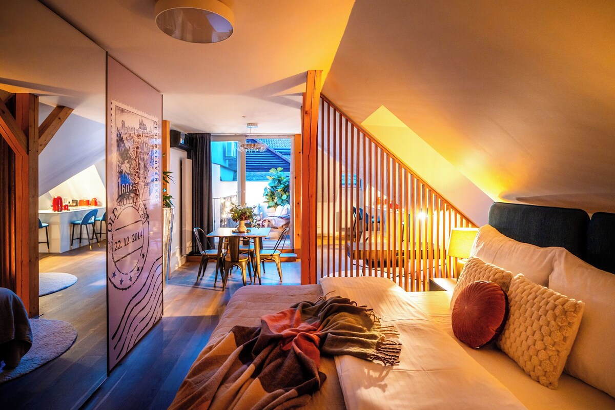 A cozy bedroom with a bed, pillows, and a blanket. There is a wooden staircase leading to an upper level. A dining area with chairs and a table is visible in the background.