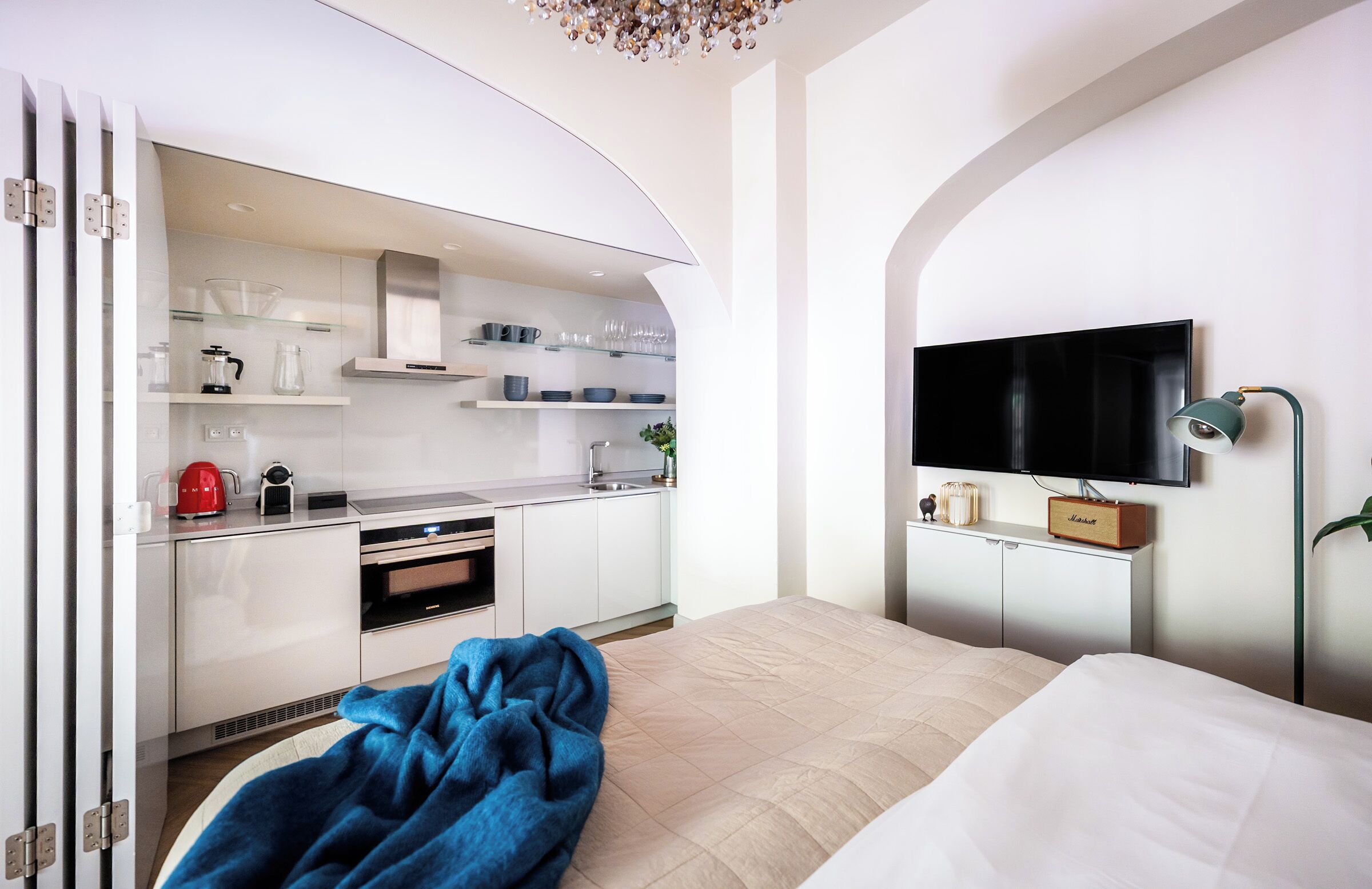 Quarto de hotel moderno com cozinha compacta, televisão de tela plana e cama com manta azul.
