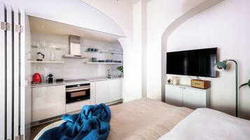 Quarto de hotel moderno com cozinha compacta, televisão de tela plana e cama com manta azul.