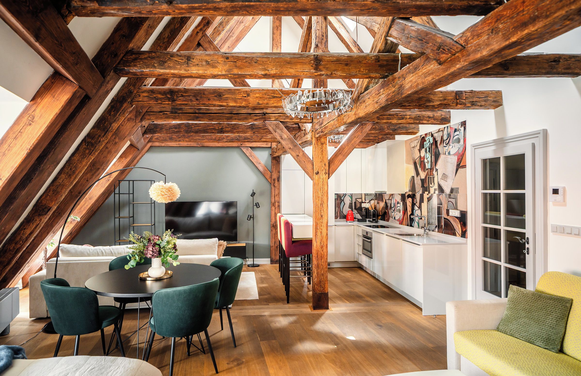 An attic room with exposed wooden beams, a dining area with a round table and chairs, a sofa, and a kitchen with a modern design.
